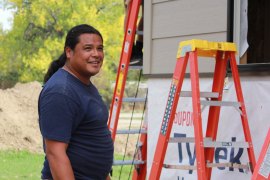 A man with long dark hair pulled into a ponytail and wearing a dark blue t-shirt stands near a building under construction.