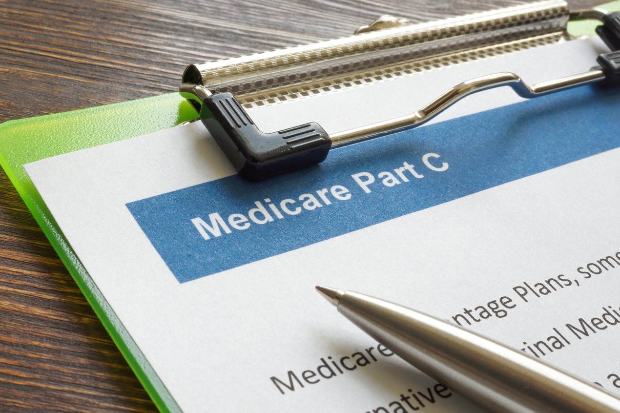 A green clipboard with a Medicare Part C form. A pen rests on top of the form.