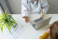 A doctor conversing with a patient. The doctor has a laptop in front of him.