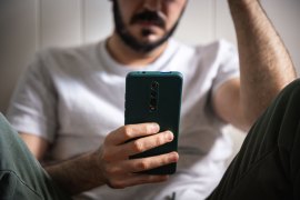 A man with a beard and wearing jeans and a t-shirt sits on the floor and looks at a phone in his hand.