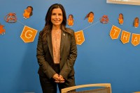 A photo of Martha Santana-Chin smiling in front of a wall painted blue. The wall appears decorated for Thanksgiving: a string of pennants spells out "Give Thanks." Images of employees' heads are superimposed on turkeys.