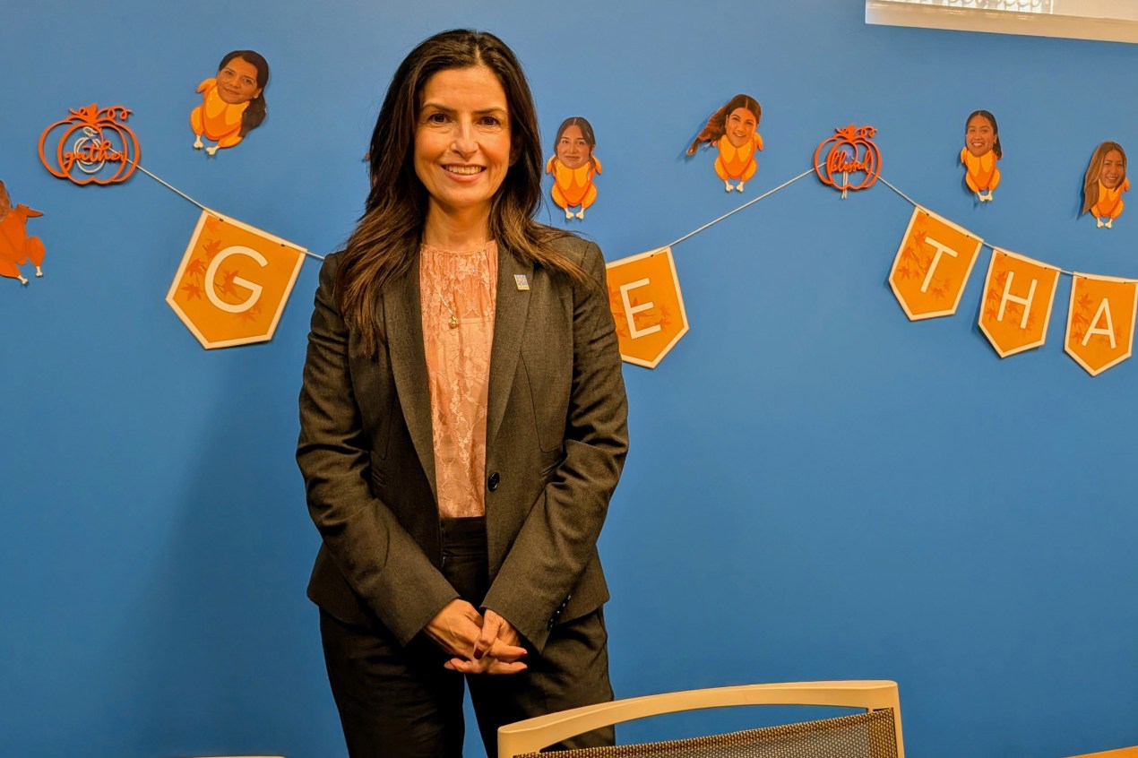 A photo of Martha Santana-Chin smiling in front of a wall painted blue. The wall appears decorated for Thanksgiving: a string of pennants spells out "Give Thanks." Images of employees' heads are superimposed on turkeys.