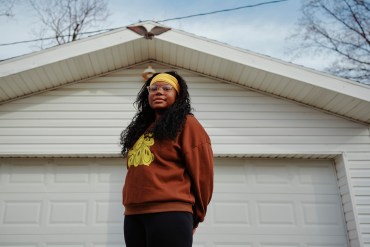 A woman wearing a yellow headband and a brown sweatshirt stands in front of a garage door, posing for a portrait.