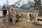 A man and a woman lean against the fences of a fenced-in area with straw on the ground and four visible goats. The woman with straight dark hair wears a dark blue sweatshirt with striped pants and smiles at the camera. The man with a beard wears a straw hat, camouflage sweatshirt, and camouflage pants is in the middle of talking and looks a something off-camera.