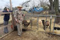 A man and a woman lean against the fences of a fenced-in area with straw on the ground and four visible goats. The woman with straight dark hair wears a dark blue sweatshirt with striped pants and smiles at the camera. The man with a beard wears a straw hat, camouflage sweatshirt, and camouflage pants is in the middle of talking and looks a something off-camera.