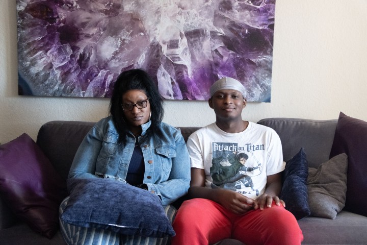 A man wearing a white t-shirt and red pants sits on a couch next to a woman wearing glasses and a denim jacket