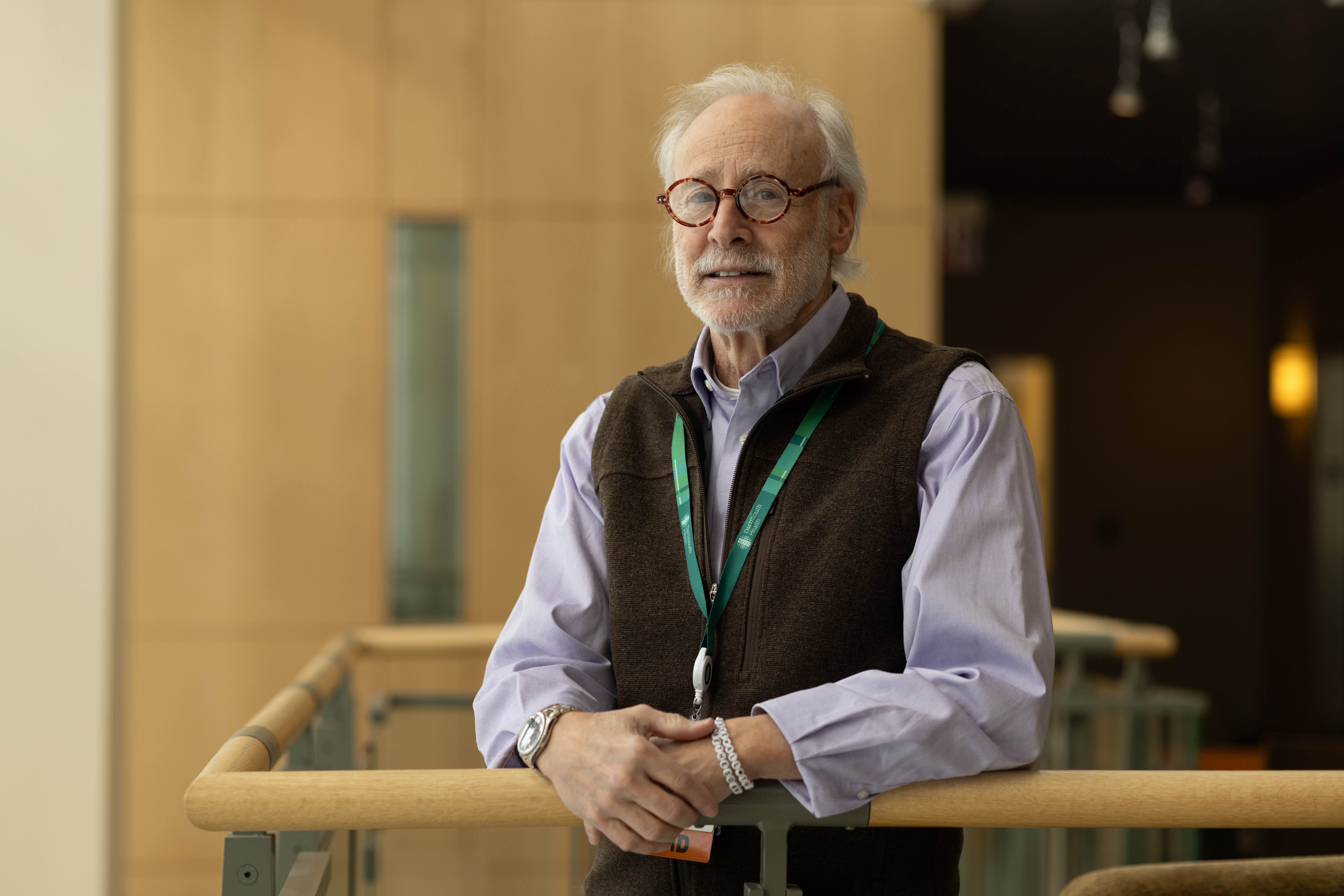 An older man in a shirt, vest and glasses leans on a rail