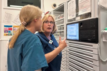 Two nurses in scrubs converse in front of a medication dispensing machine at a hospital.