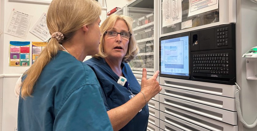 Two nurses in scrubs converse in front of a medication dispensing machine at a hospital.