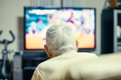 An older man is seen from behind lounging in a chair. A TV is on in front of him.