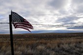 An American flag blows in the wind next to a barbed-wire fence in front of a landscape of grasslands
