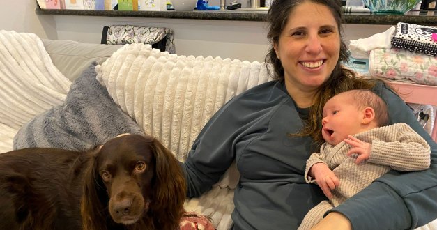 A woman sits on a couch with a young baby in her left arm and her right hand petting a brown dog sitting beside her. She smiles at the camera.