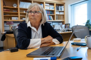 A researcher sits at her desk indoors. A laptop is in front of her.