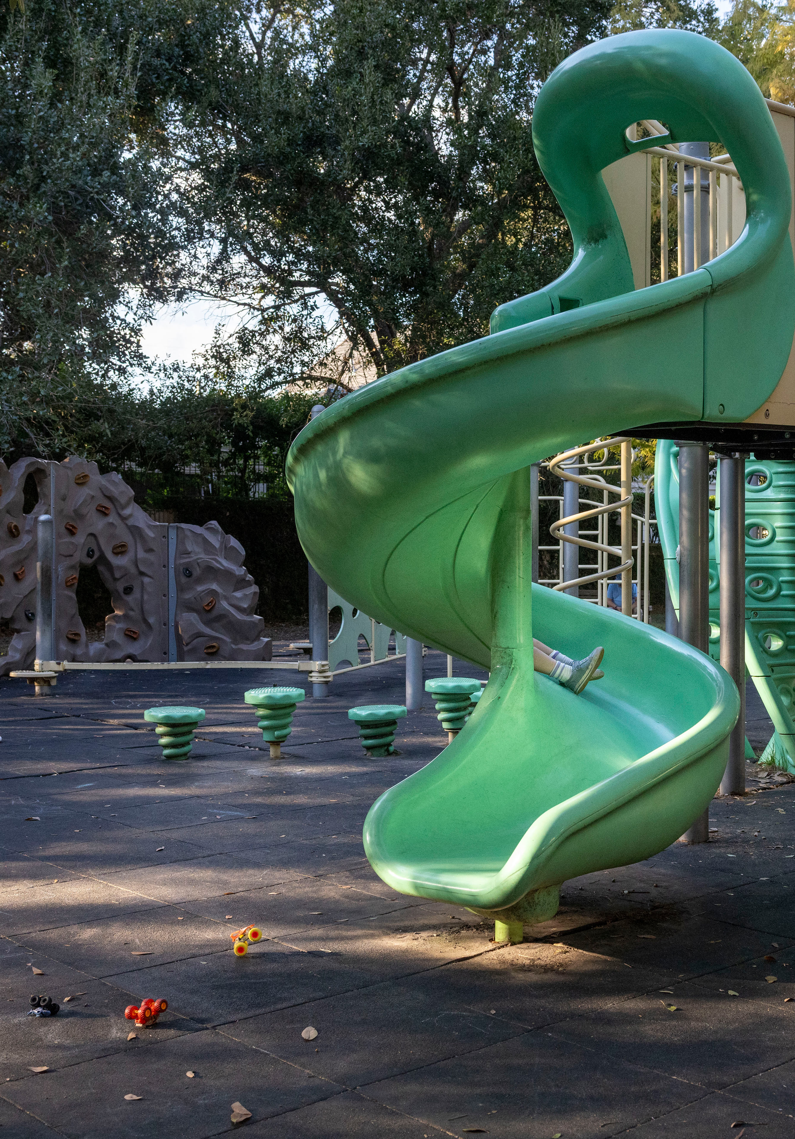 A child's feet can be seen sticking out from one of the twists of a green plastic slide at a playground.