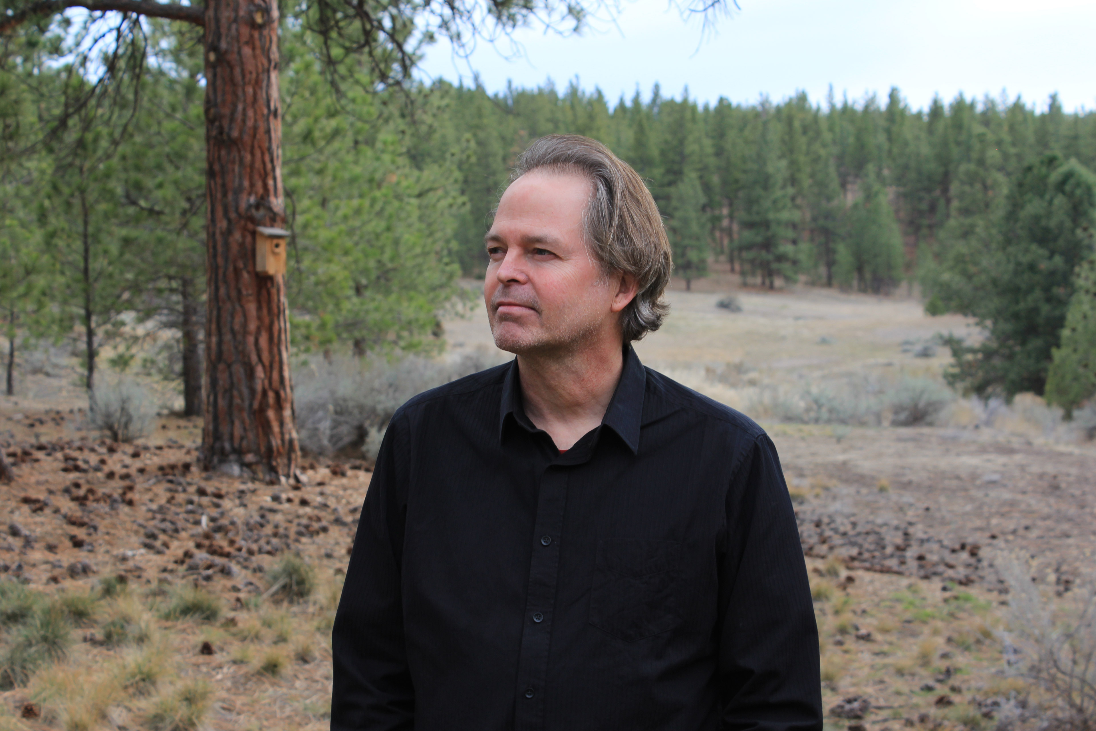 Philip Stewart stands outside in a wooded area. Evergreen trees are seen behind him.