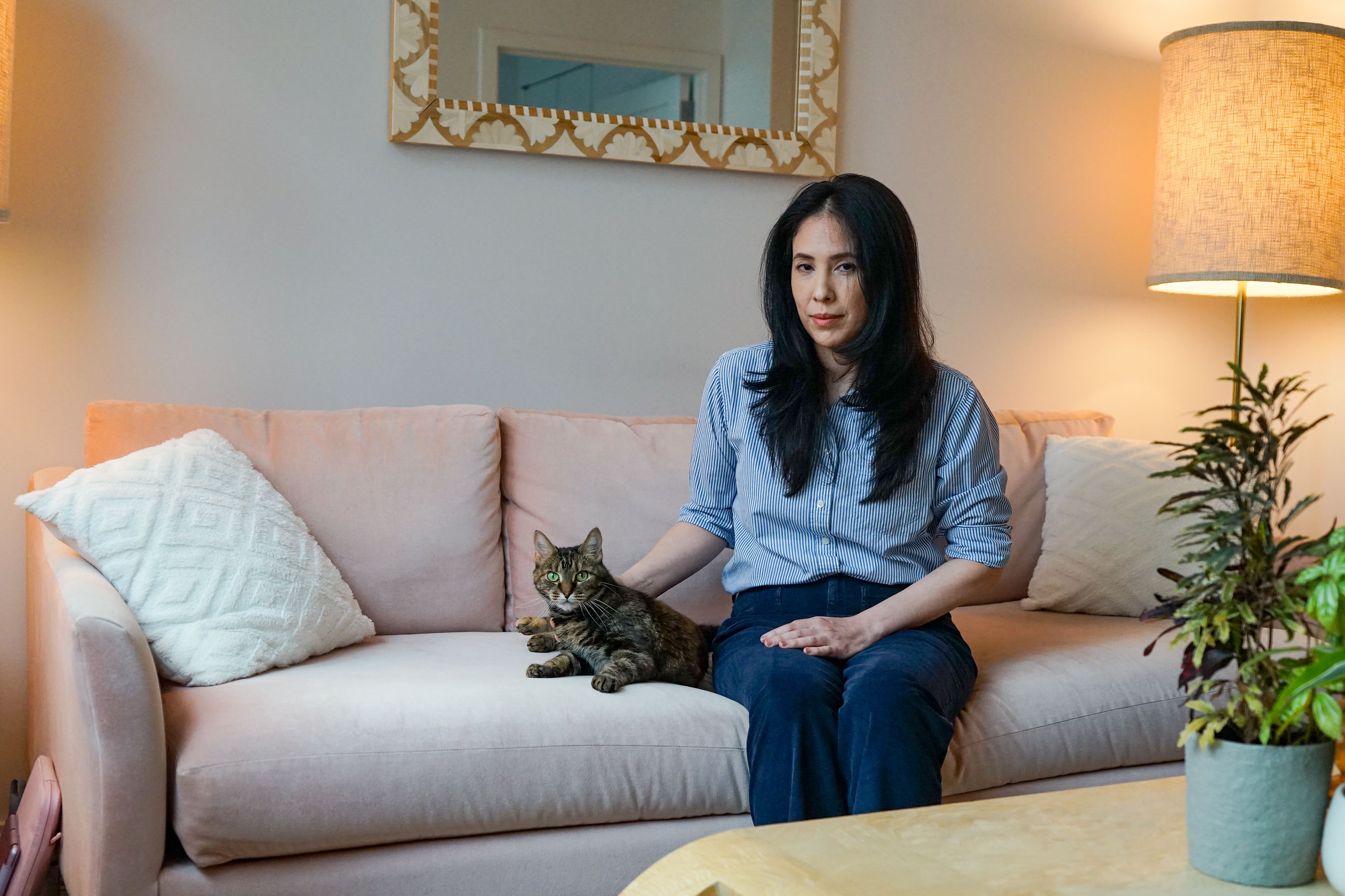 A woman sits on a couch with a tabby cat beside her. Her hand gently rests beside her cat.