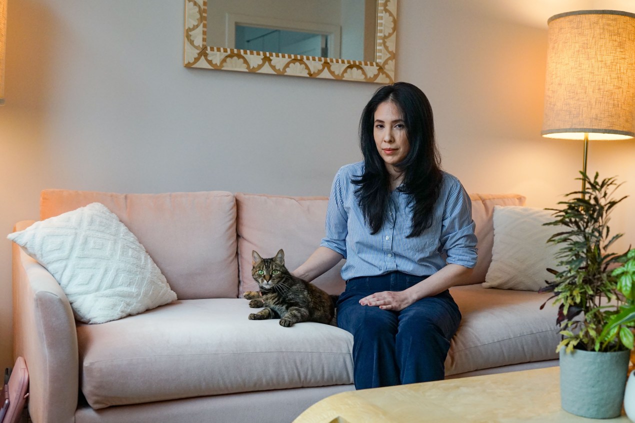 A woman sits on a couch with a tabby cat beside her. Her hand gently rests beside her cat.