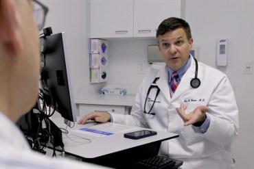 A male doctor in a white coak speaks to someone out of frame.