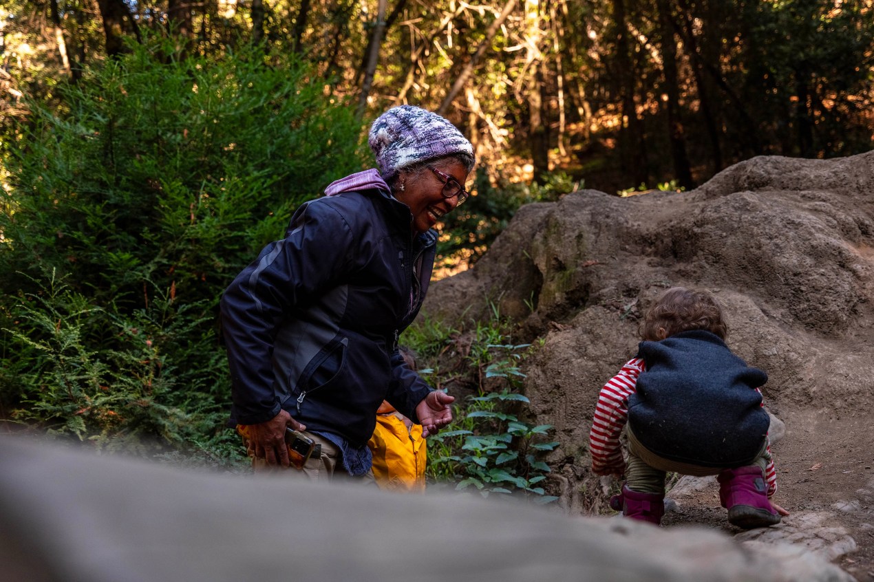 A senior woman in active outdoor clothing encourages a toddler to navigate uneven ground in a lush forest environment.