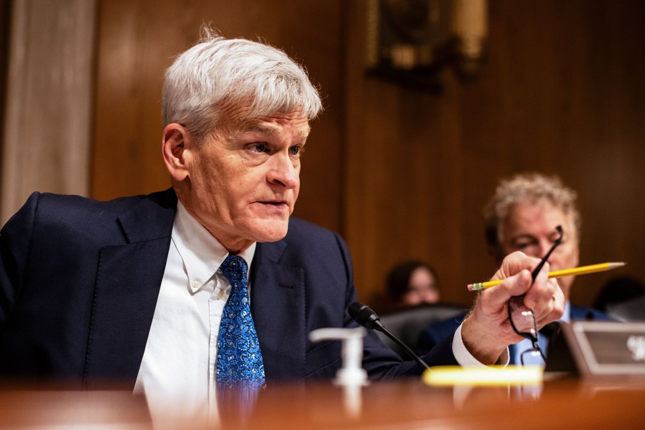 Bill Cassidy sits behind the dais in a Senate hearing room. He is speaking while holding both his glasses and a pencil in his left hand.