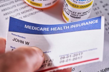 A man holds a copy of his Medicare card. Orange pill bottles and medical forms are seen behind it.