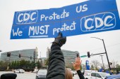 A hand holds up a sign next to traffic in an intersection that says "CDC protects us, we must protect CDC"