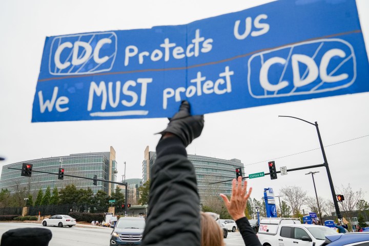 A hand holds up a sign next to traffic in an intersection that says "CDC protects us, we must protect CDC"