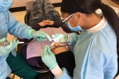 A photo from above of a person lying in a dental chair having work done. A dentist and assistant sit on either side of the patient's head.