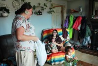A doula stands next to a young mother sitting in a chair draped with colorful fabric. She holds a young baby in her arms.