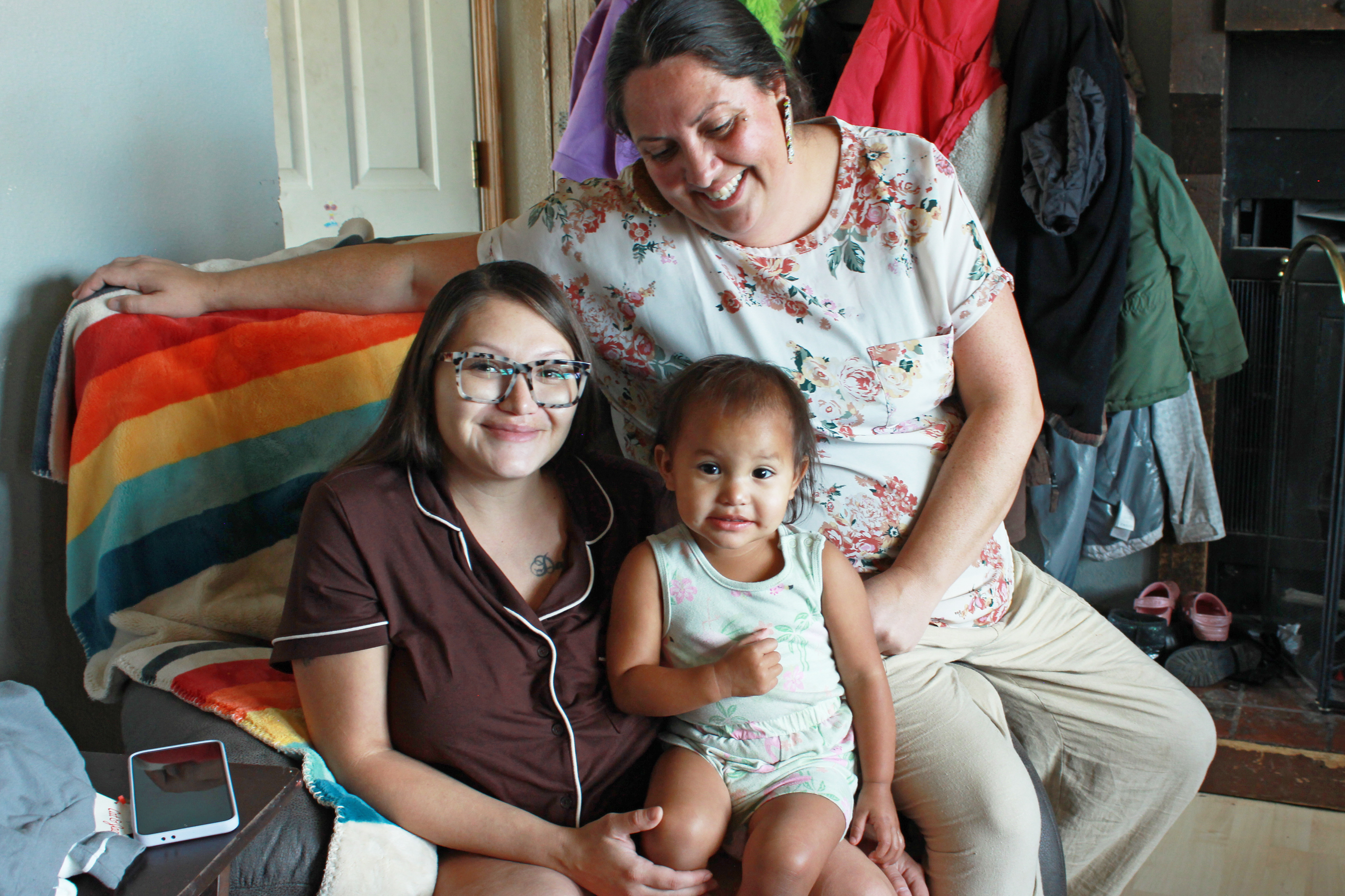 Britney WolfVoice sits in a chair draped with a rainbow-colored blanket. Her daughter Ellie sits in her lap. Misty Pipe is seated behind them. All three are smiling.
