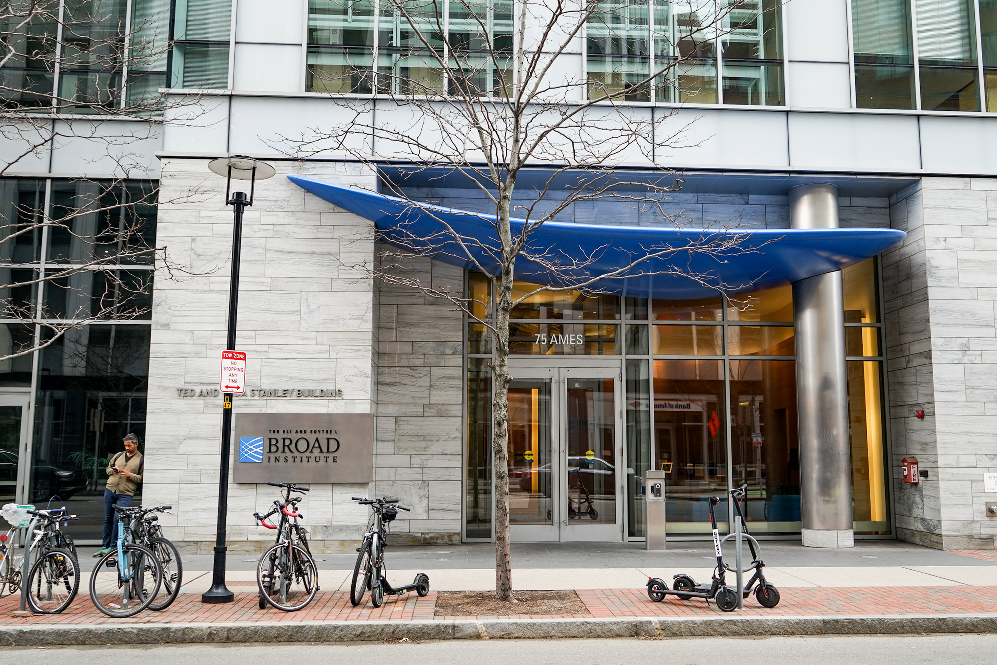 An exterior shot of a large building. A sign on the outside reads, "The Eli and Edythel Broad Institute." A traffic island in front of the building has bikes and electric scooters parked in front of it.