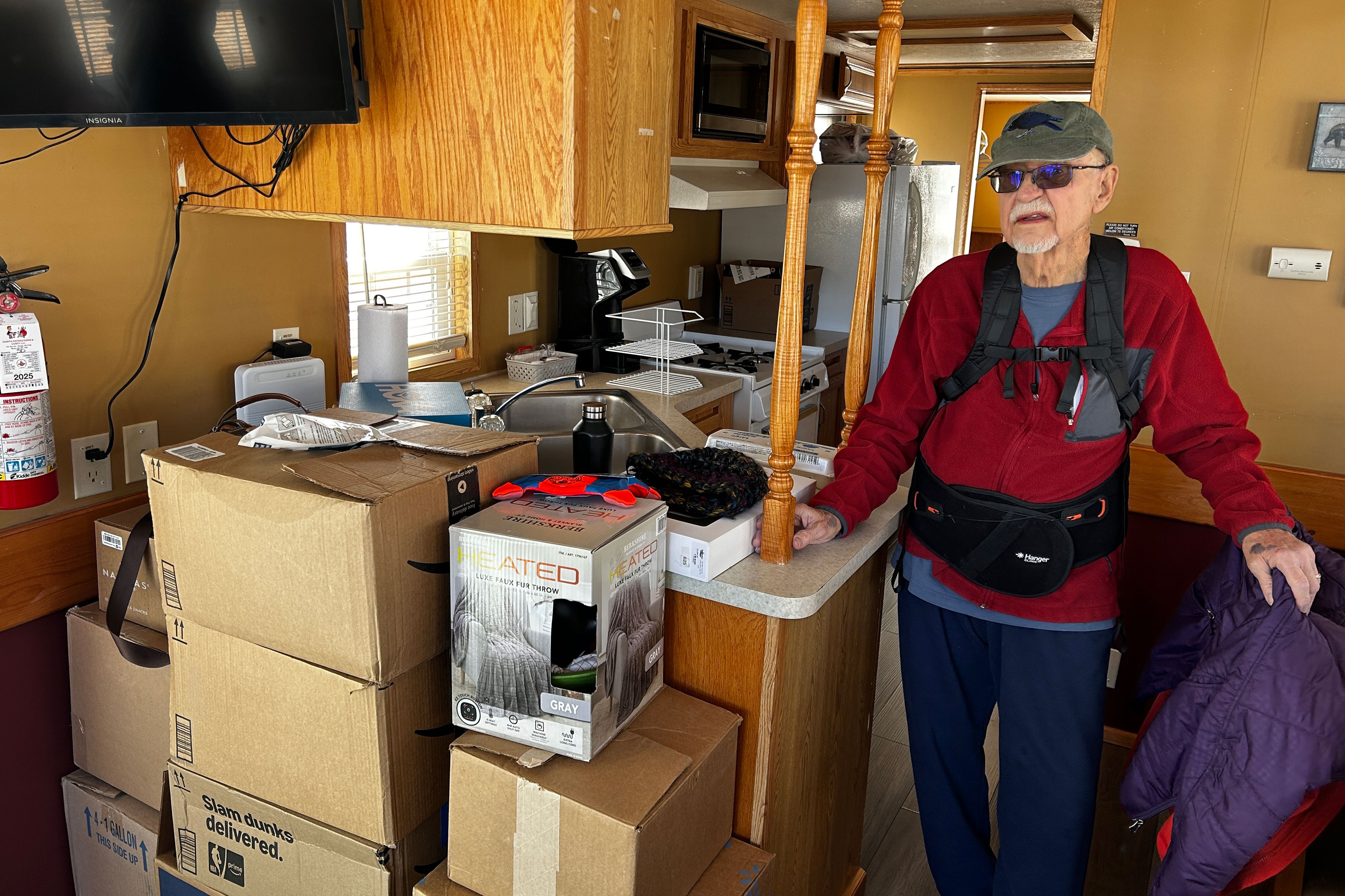 An older man stands indoors next to a pile of packed cardboard boxes.