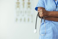 An unidentifiable medical professional stands to the side of frame in blue scrubs and holds a stethoscope.