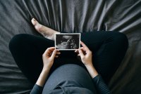 A high-angle photograph of a pregnant person sitting cross-legged on a bed and holds a sonogram of a baby.