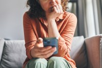 A woman sitting on a couch indoors, appearing focused and concerned while looking at her smartphone.