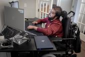 A man with muscular dystrophy works at a desk with multiple computer monitors.