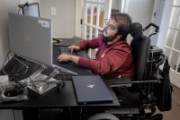 A man with muscular dystrophy works at a desk with multiple computer monitors.