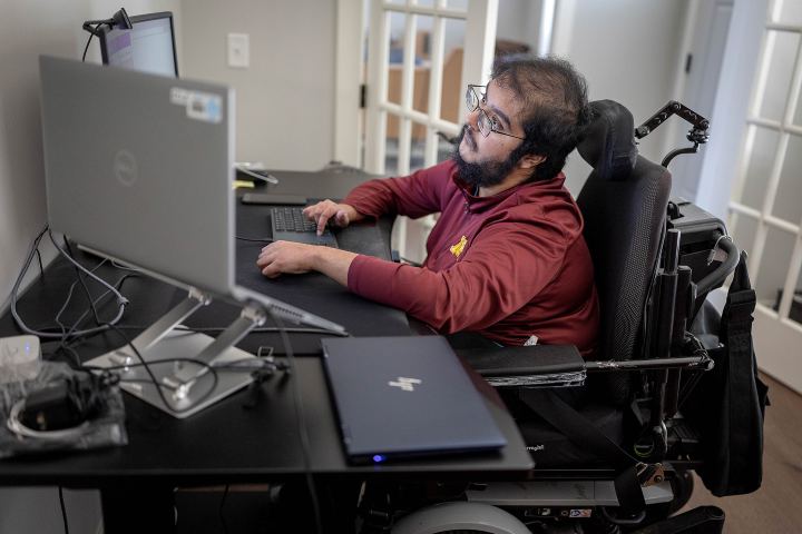A man with muscular dystrophy works at a desk with multiple computer monitors.