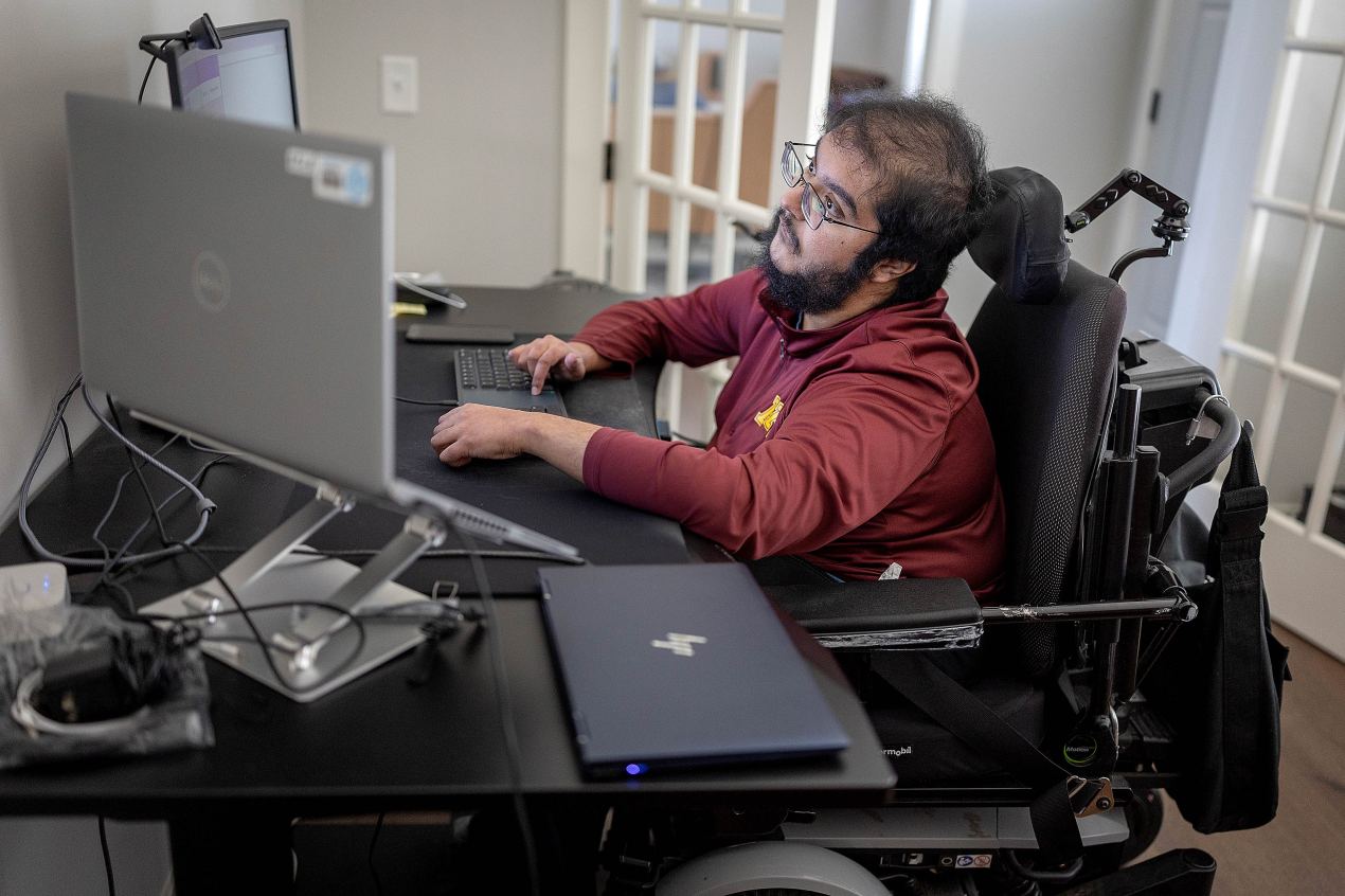A man with muscular dystrophy works at a desk with multiple computer monitors.