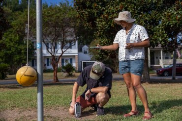 Two adults work outside. One on the left uses a device pressed to the ground to test for lead. Another holds out her arm, phone in hand, to get coordinates.