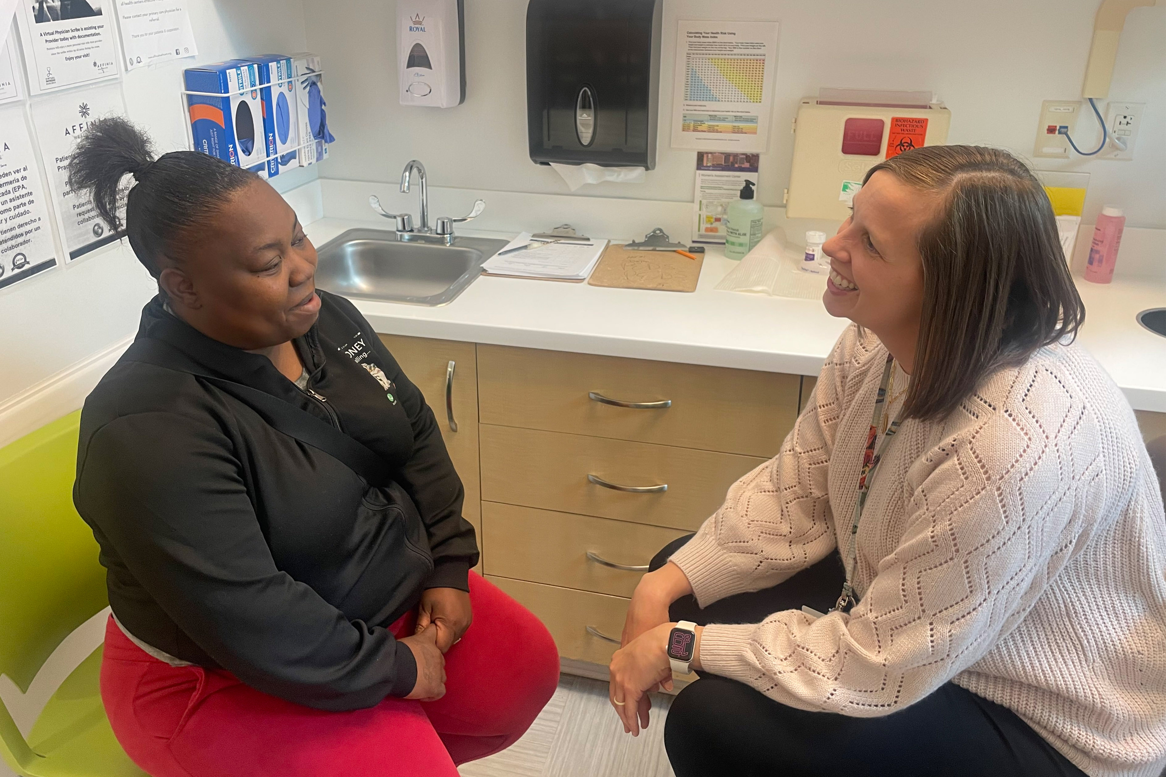 A doctor sits on the right, speaking to her patient, seated on the left side of the frame.