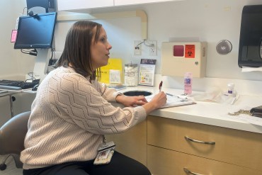 A doctor writes notes in an examination room while talking to a patient out of frame.