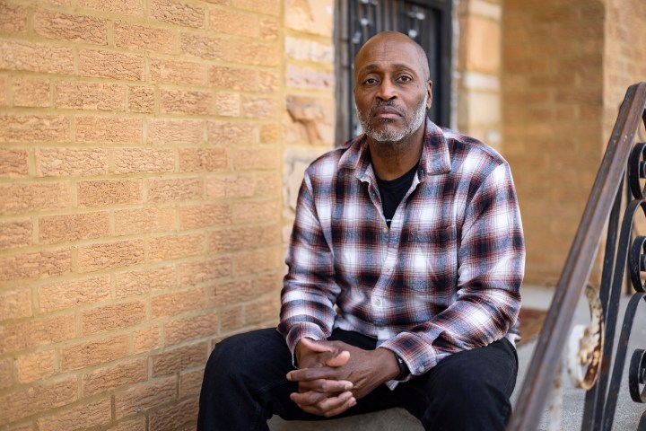 A man wearing a plaid button-up shirt sits on the front step of a home for a portrait