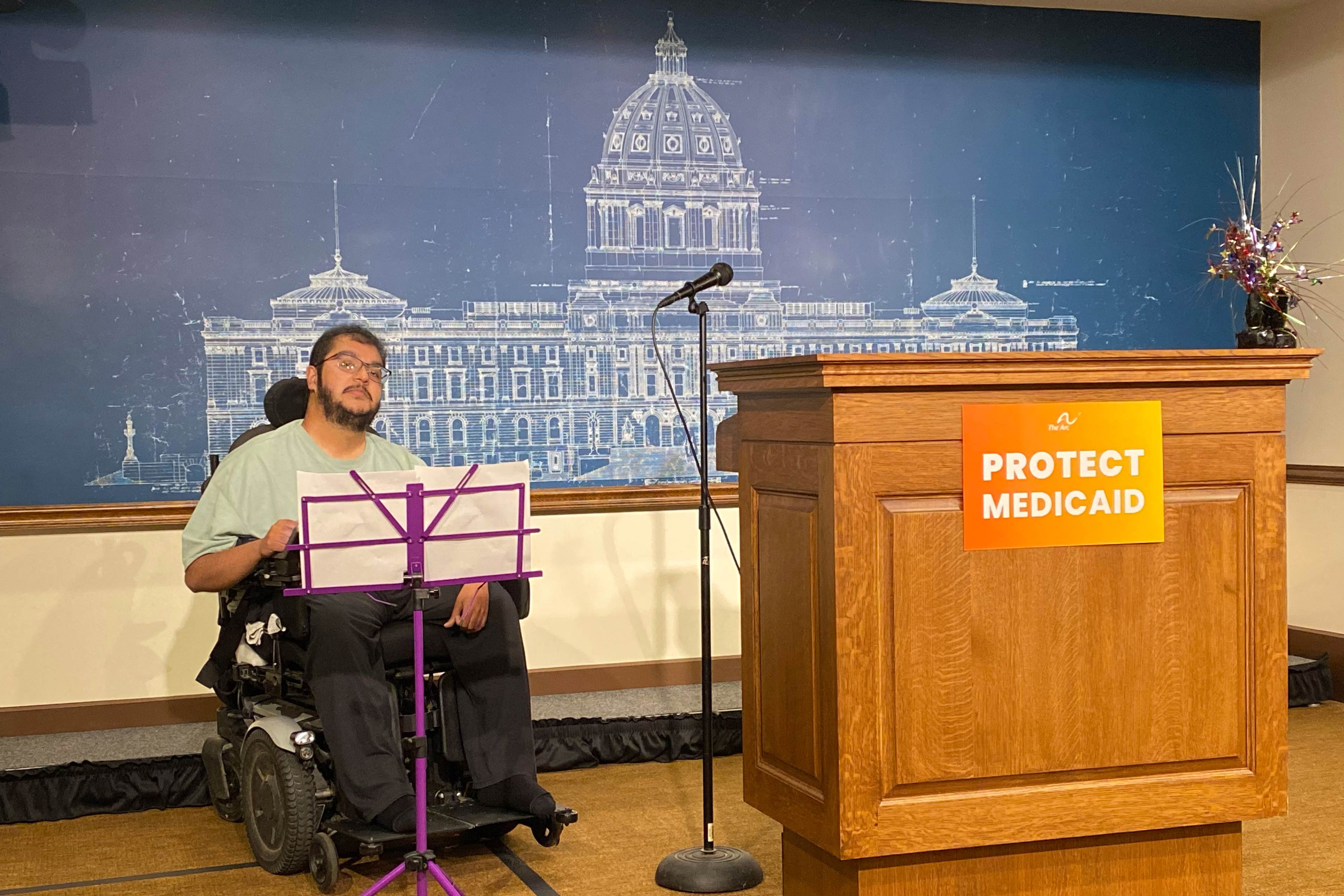 Sumukha Terakanambi, a man with muscular dystrophy, is on a stage sitting in a powered wheelchair. Next to him is a podium with a sign that reads, "Protect Medicaid."