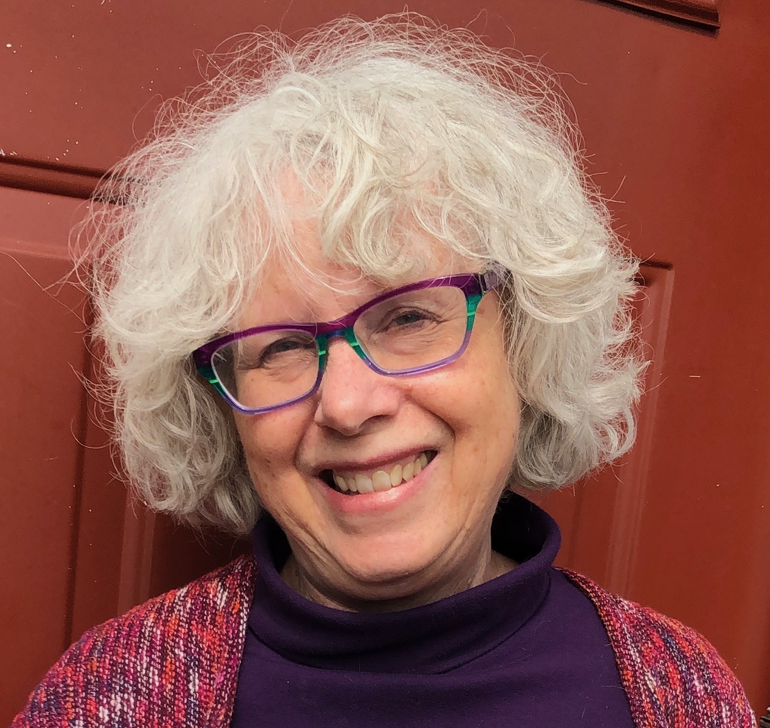Beth Spencer — a recently retired social worker — smiles for the camera. She has gray wavy hair and colorful glasses.