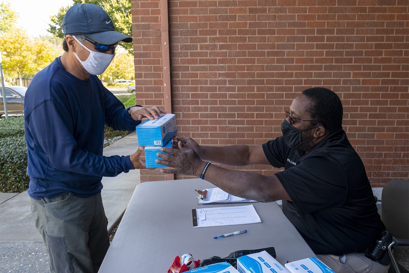 Kemper Williams sits behind a table on the right, handing boxes of masks to Son Chong on the left. Both are wearing masks.