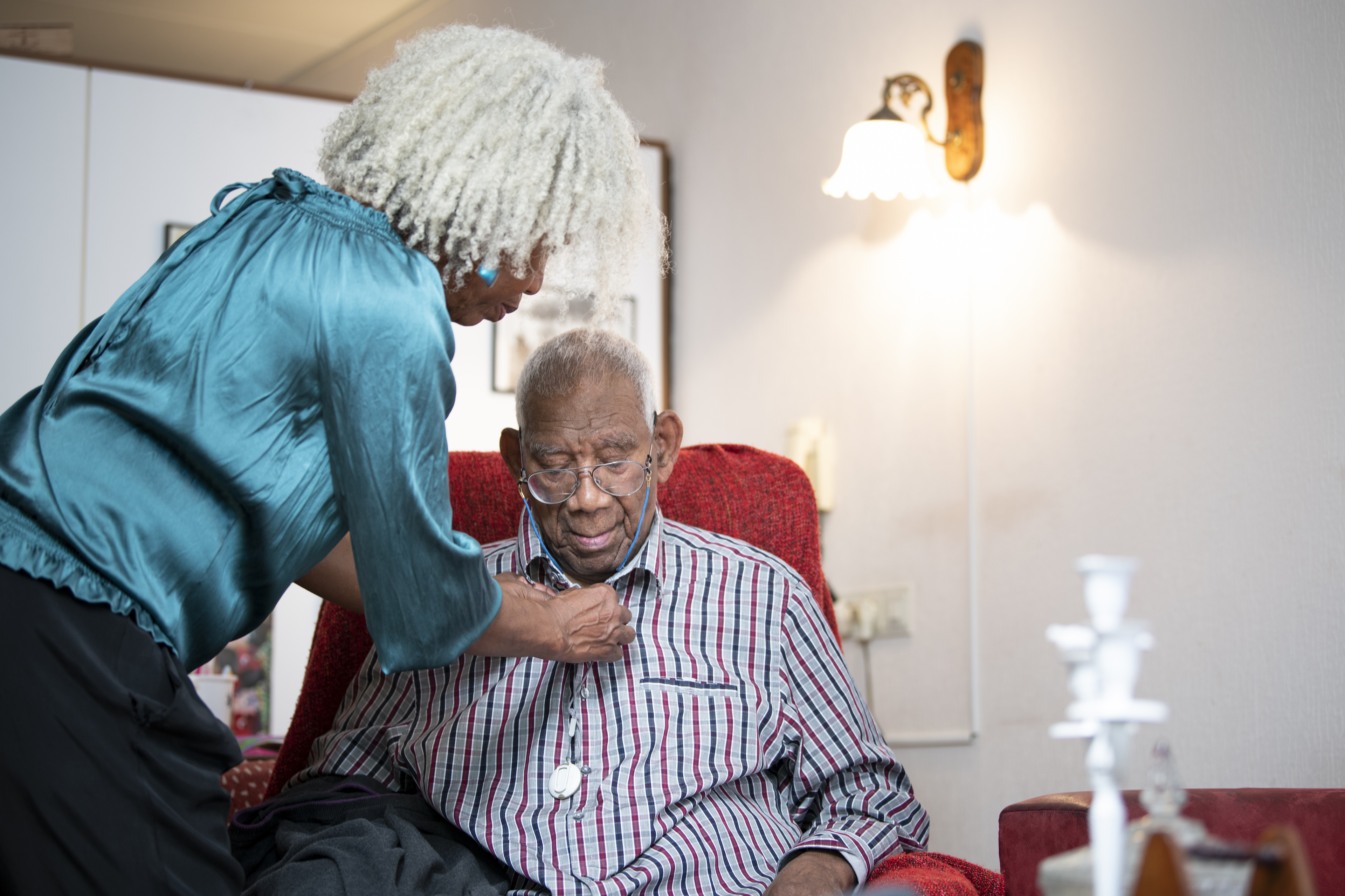 On the left side of the image, a Black woman with short, white dreadlocks leans over a senior man, her father, to help button his shirt. The woman wears a turquoise shirt, while the man sits in a red recliner. They are in their home living room.