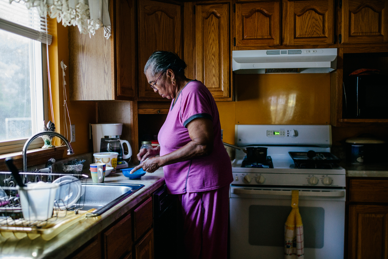 A senior black woman is standing in a kitchen making breakfast for herself. She is seen in profile facing the left towards the kitchen window.
