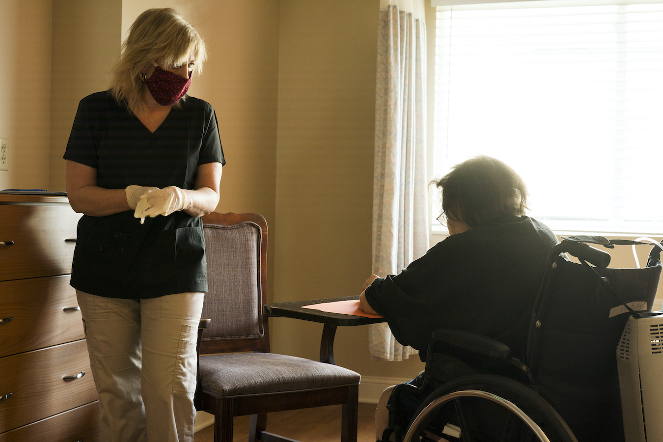 Blond female healthcare worker wearing scrubs, gloves and a red fabric face mask approaches a patient on oxygen sitting in a wheelchair to begin a speech therapy session amid the corona virus COVID-19 pandemic.