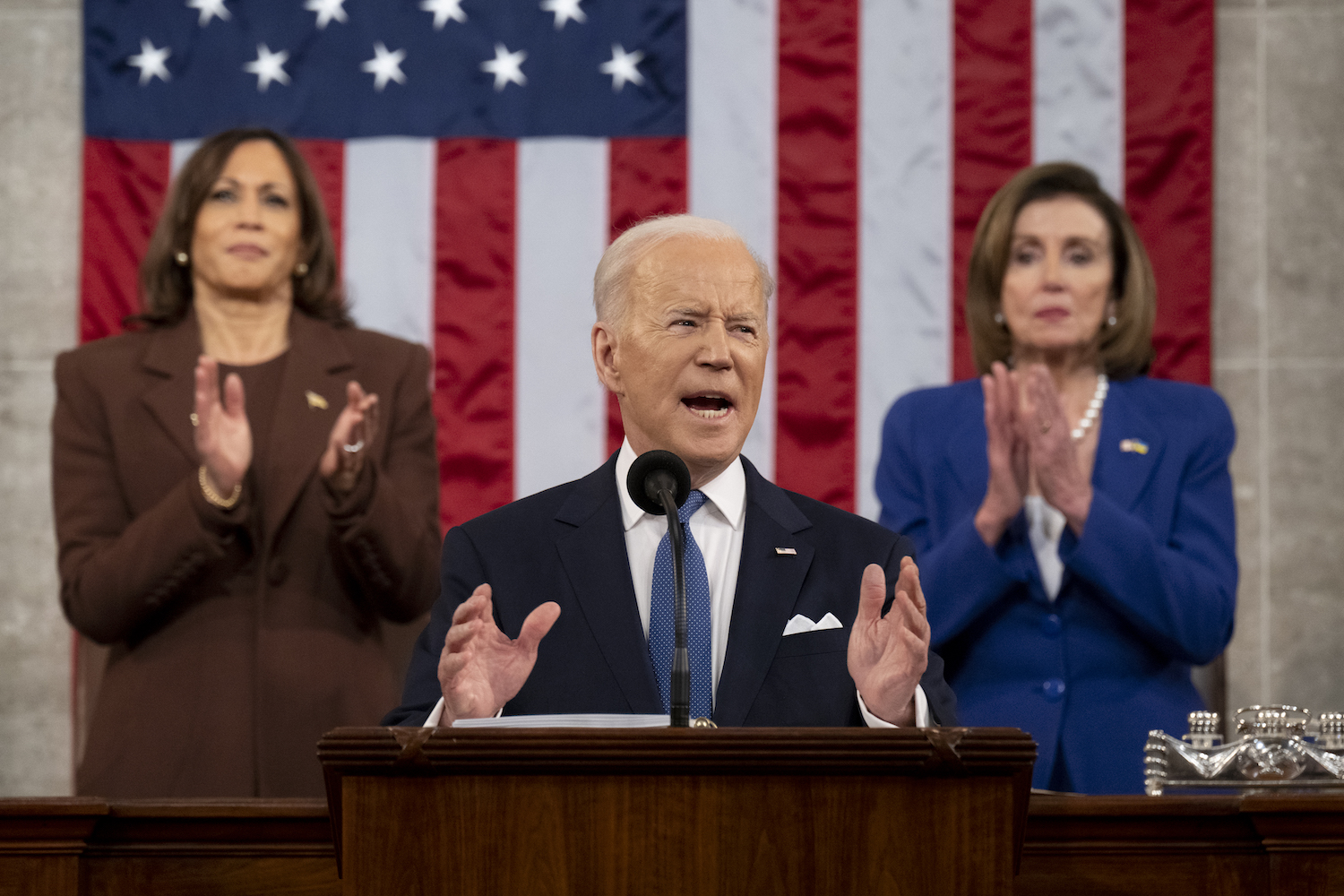 President Joe Biden speaks from a podium with Vice President Kamala Harris and House Speaker Nancy Pelosi standing behind him.